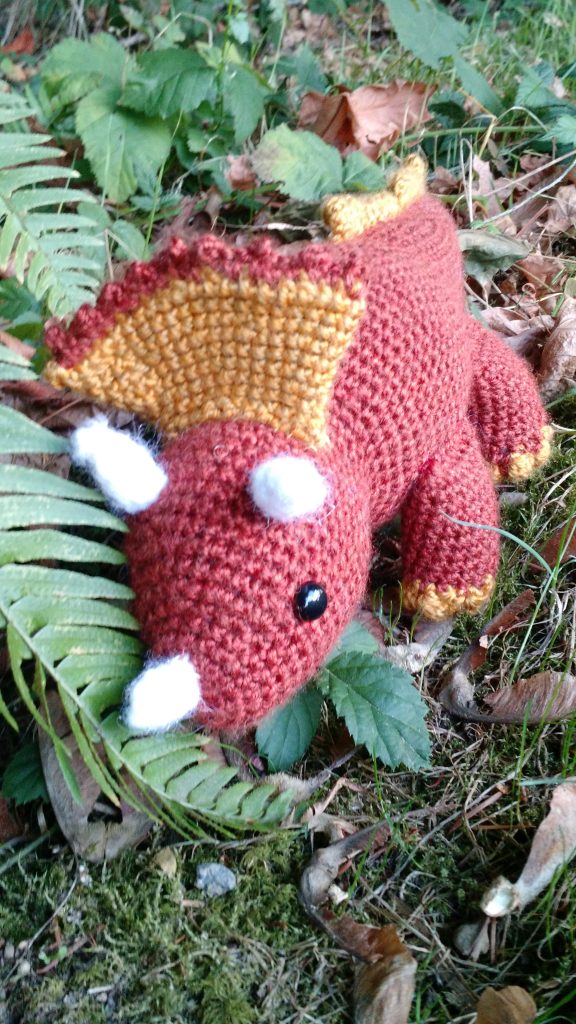 Photo of a crocheted stuffed toy Triceratops nestled in some ferns. It is red with a yellow crest.